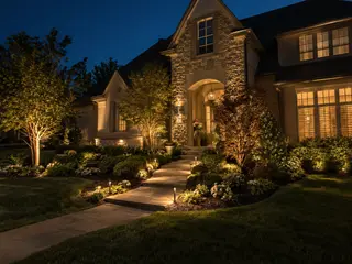 Warm uplighting highlighting the stone facade, trees, and walkway of a Johnson County home at dusk