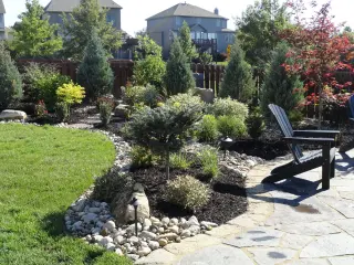 Layered evergreen and Japanese maple plantings around a river rock bed beside a flagstone patio